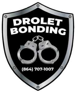 The Drolet Bail Bonds logo, commonly seen by fans as bearing a striking resemblance to the old Oakland Raiders logo, has Drolet Bonding text across the top of the image, while it has silver handcuffs with different gradients and reflections in the middle of the shield, and the company phone number is located at the bottom of the shield. That number reads (864) 707-1007. The shield logo represents our expansion to Walhalla, SC, and all surrounding Upstate counties.
