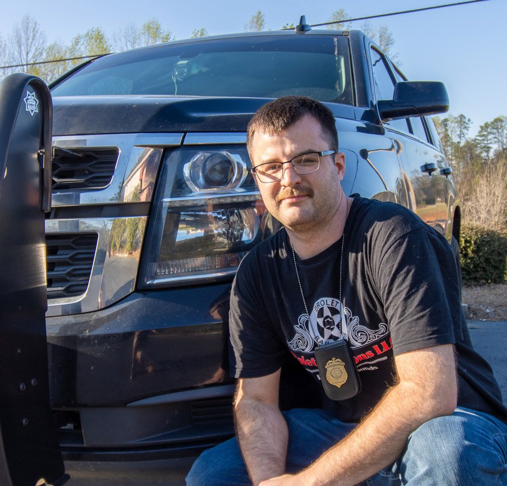 Ryan Drolet, of Drolet Bail Bonds, a family bail bond company in Walhalla, SC, poses for the camera in front of his 2019 Chevrolet Tahoe with a police push bumper attached to the front of the truck. He has a short, military-style haircut with a Drolet Bail Bonds t-shirt and a necklace badge holder with a bail bondsman/fugitive recovery agent badge attached to it.