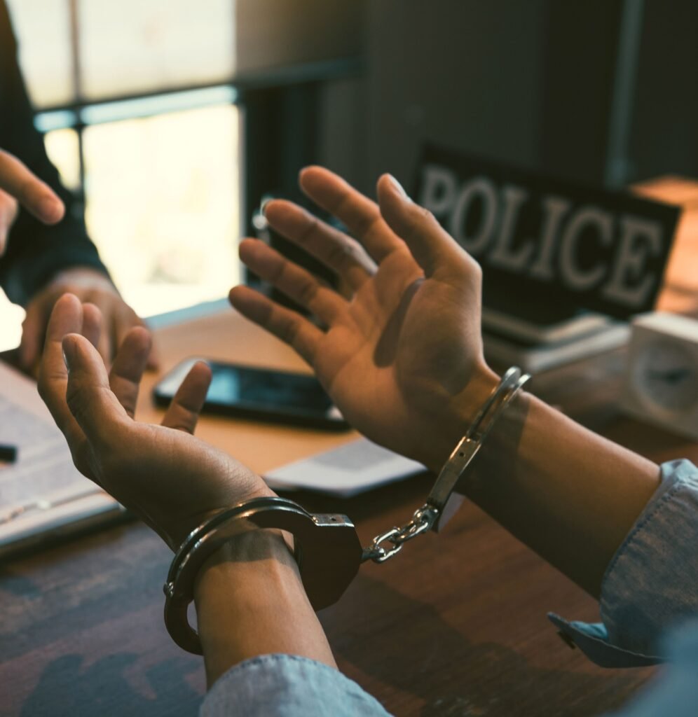 The hands of a young man are shown with handcuffs on his wrists. He is being questioned by an officer of the Walhalla Police Department over allegations that he stole lawn equipment from a local elderly man. A black sign with white letters reads "POLICE" as the cop sits across a wooden table from the defendant.